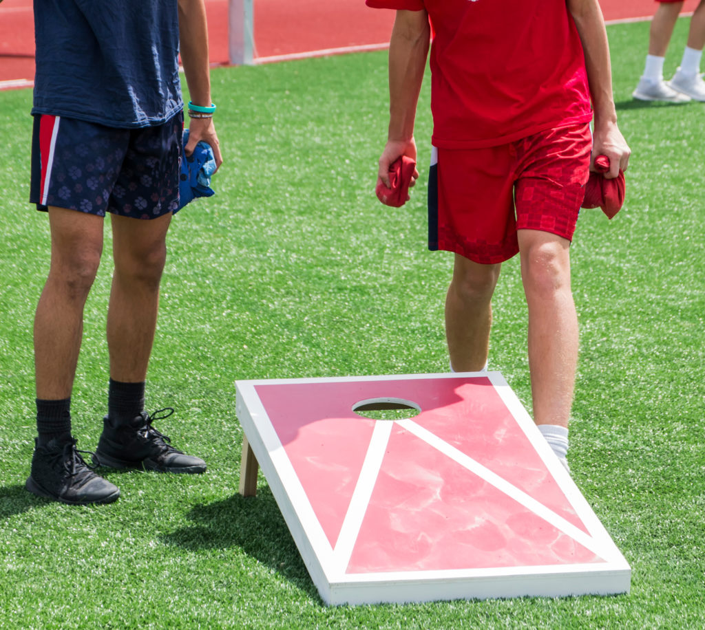 Two boys play corn hole during gym class on a green turf field in the sunshine.